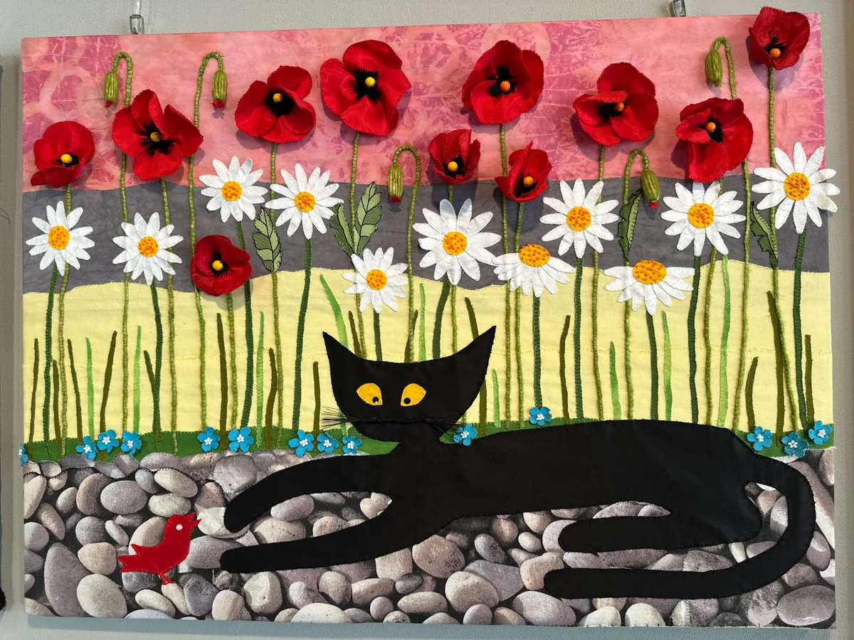 Ein Stoffbild, bei dem gleich mehrere Elemente ineinander spielen: Katze, Blumen, roter Vogel. Bild aus Stoff.